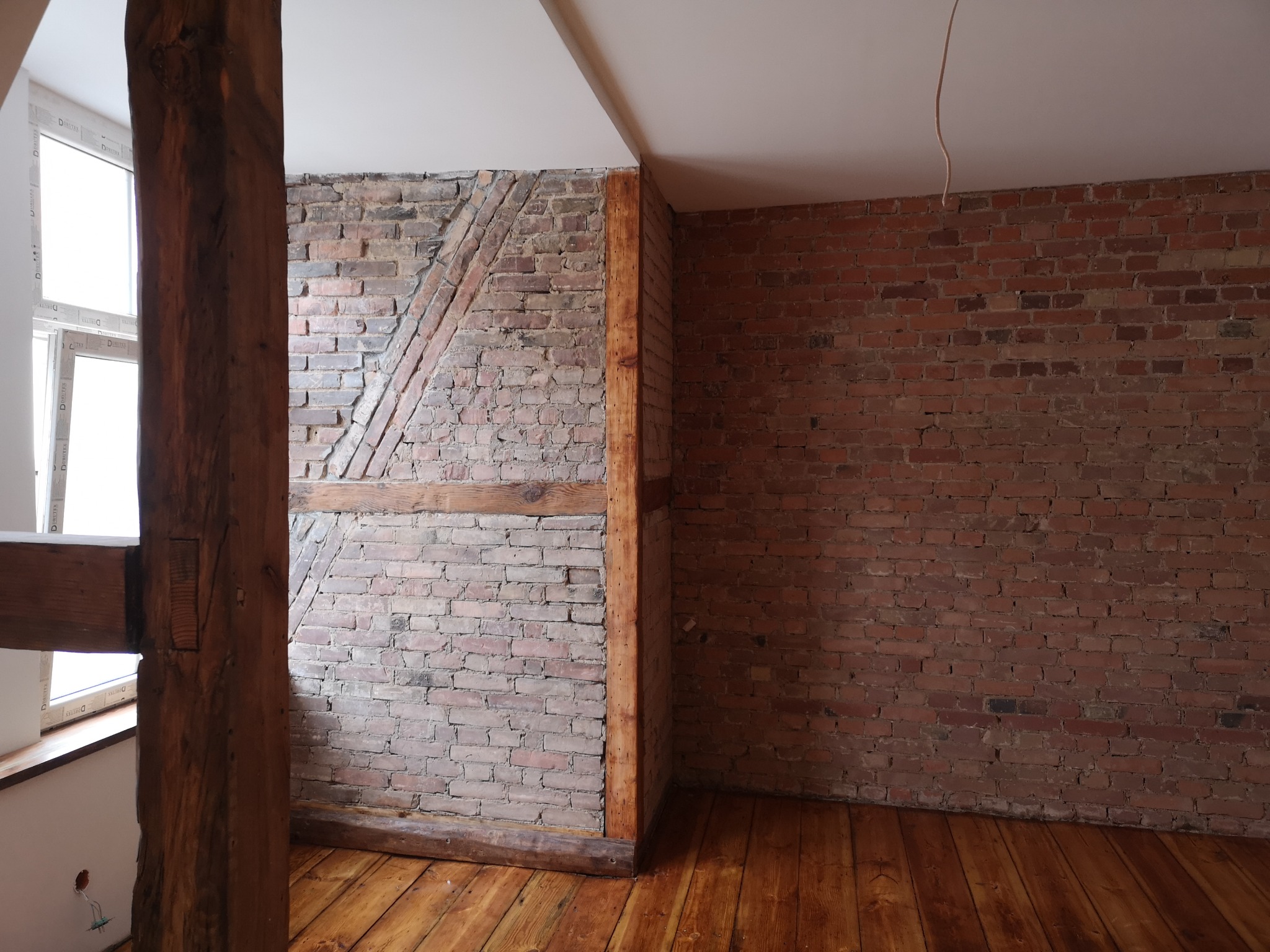 Surowe wnętrze z odsłoniętą cegłą, drewnianymi belkami i podłogą, w trakcie prac remontowych, widoczne ślady po instalacjach elektrycznych.