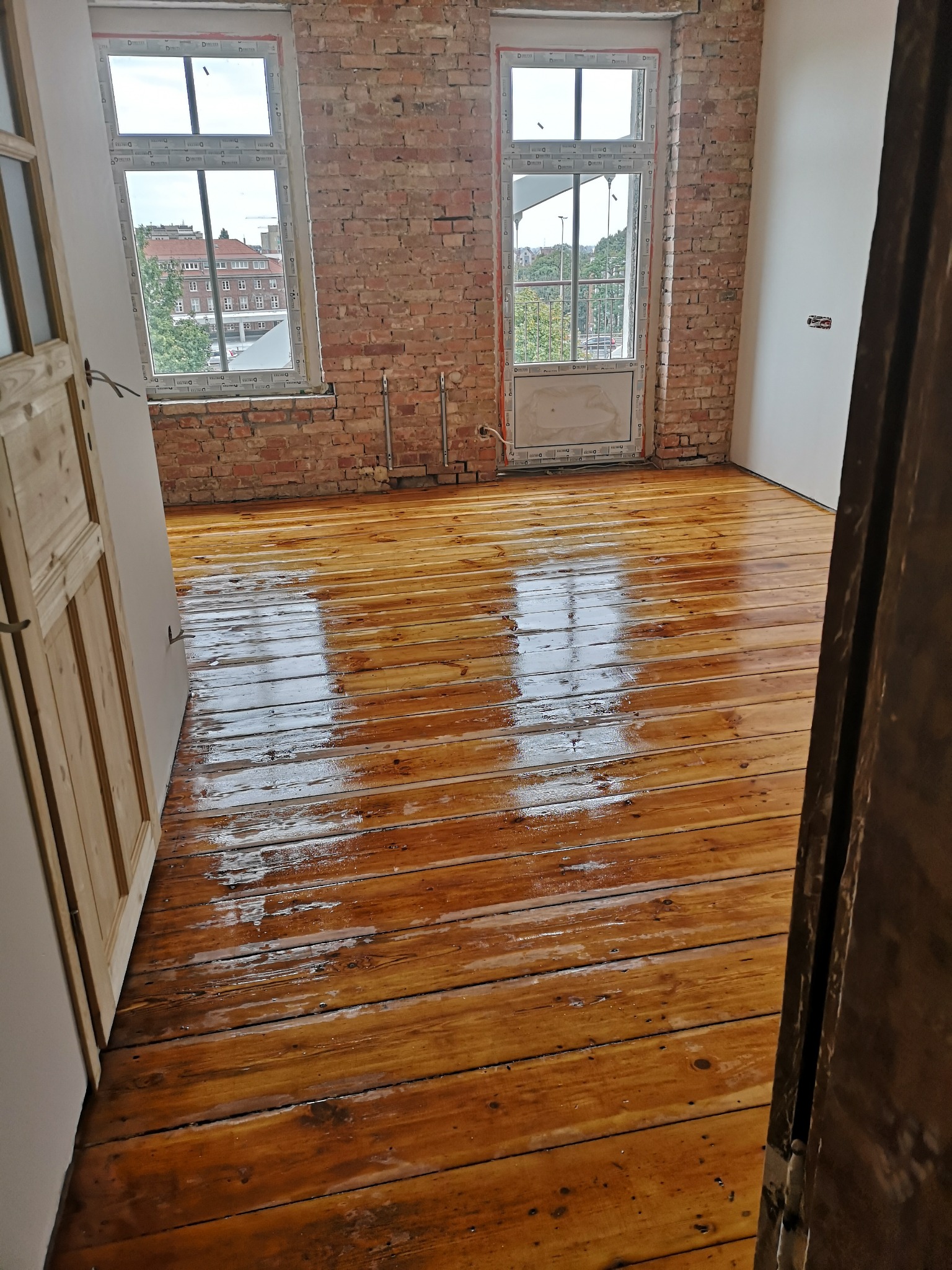 Odnowiona drewniana podłoga w pokoju z odsłoniętą cegłą na ścianie, widok przez otwarte drzwi.