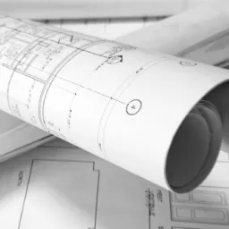 Zwinięty plan architektoniczny w czerni i bieli, leżący na rozłożonych rysunkach technicznych; widoczny fragment z oznaczeniem '4' w okręgu i wymiarami.