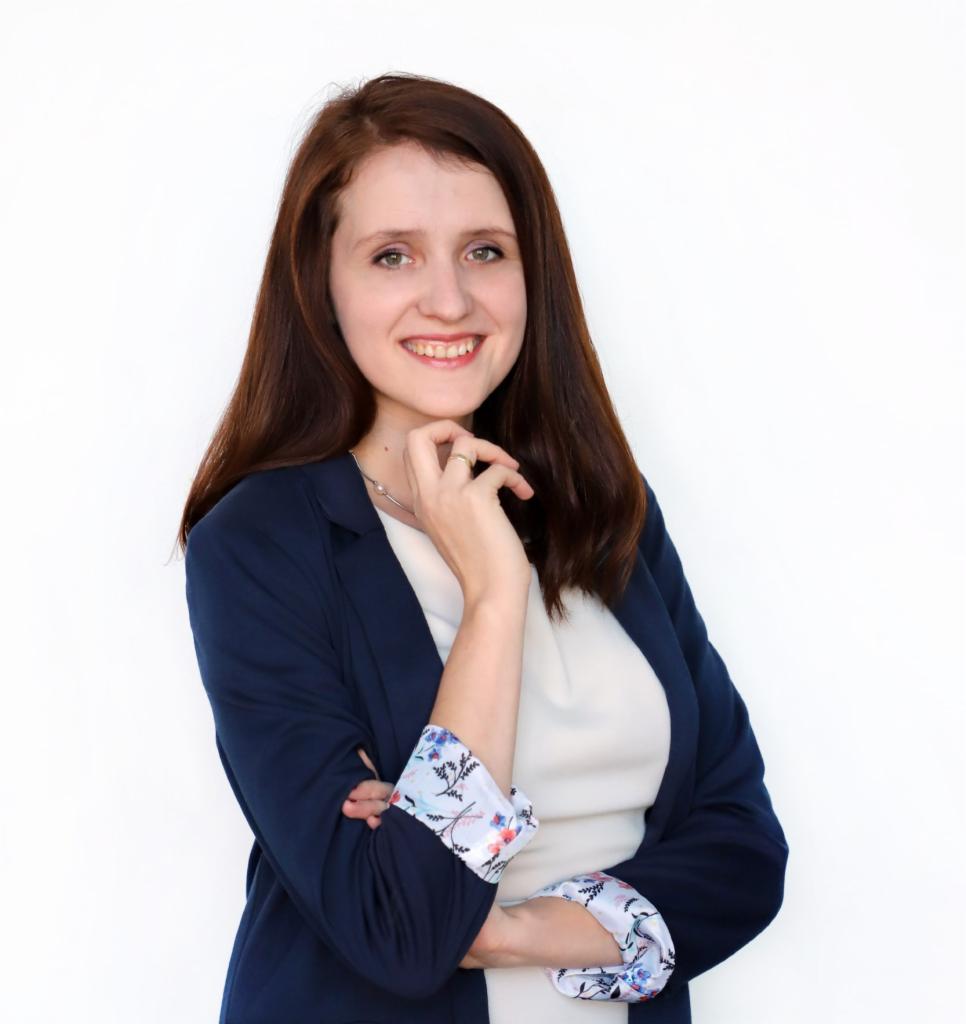 Uśmiechnięta brunetka w granatowej marynarce z wzorzystymi mankietami, opierająca brodę na dłoni, na białym tle.