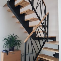 ADAM KUBALA - Ażurowe schody z drewnianymi stopniami i czarną, geometryczną balustradą, widok z dołu, obok szafka z rośliną doniczkową.