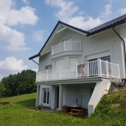 Białe balustrady balkonowe z pionowymi szczeblami na dwóch poziomach domu z szarą elewacją, widok z dołu na tle błękitnego nieba z chmurami.