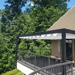 ADAM KUBALA - Zadaszony taras z czarną metalową konstrukcją i balustradą, widok na bujną zieleń drzew i dach domu z lukarną.