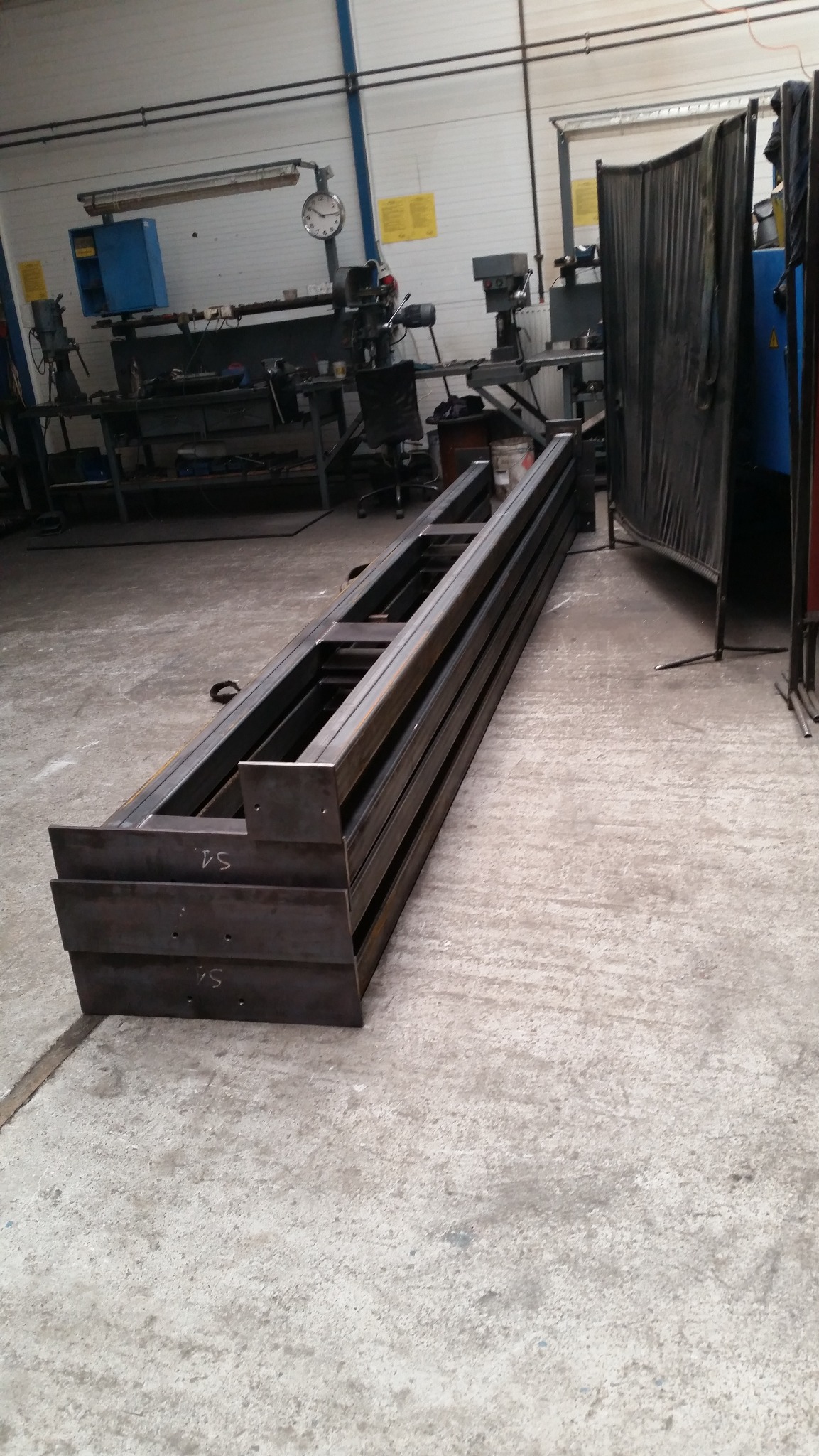 Stos belek stalowych o przekroju prostokątnym, z widocznymi śladami spawania, leżący na betonowej podłodze w warsztacie, w tle maszyny i narzędzia.