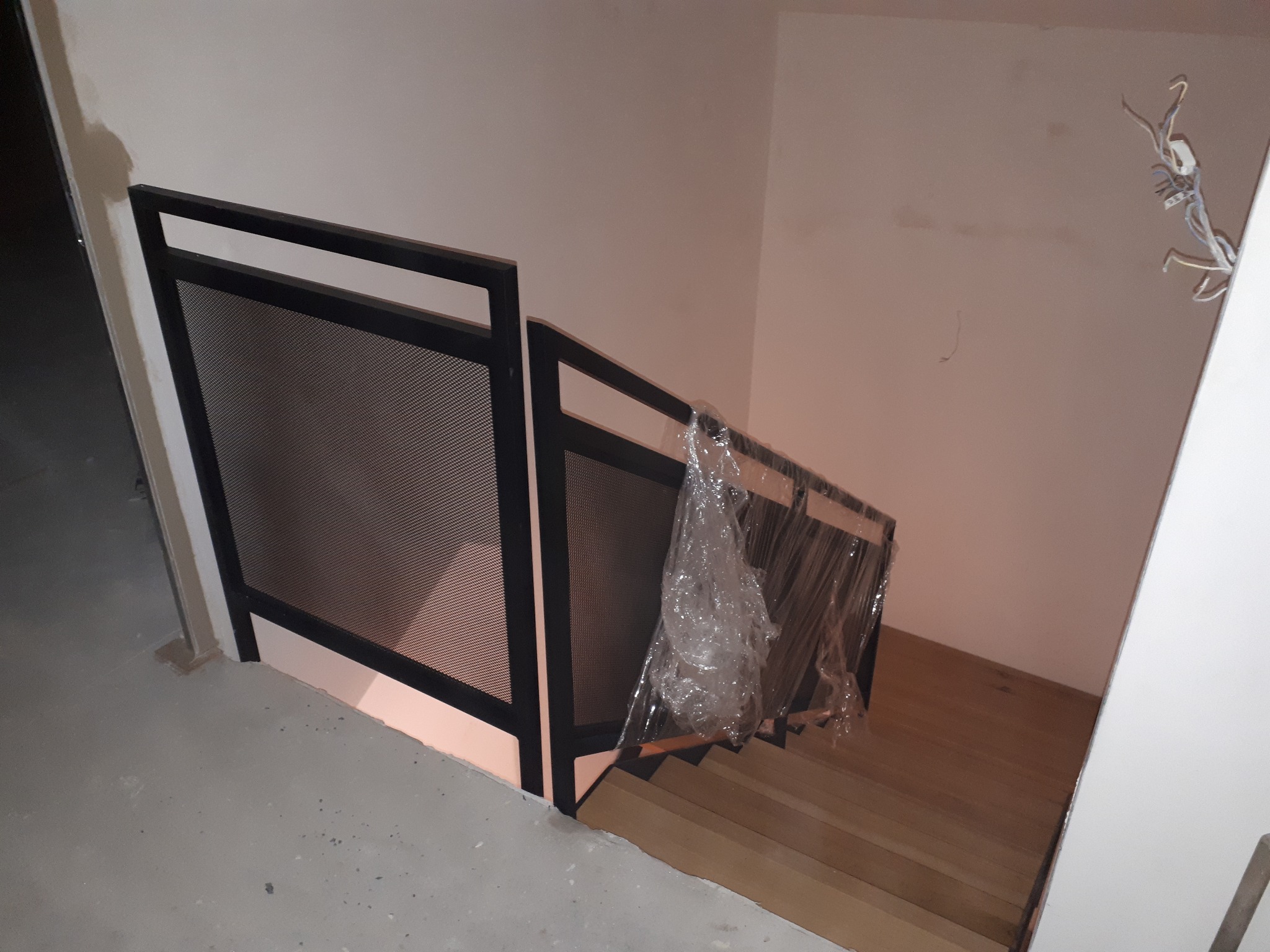 Nowoczesne schody z drewnianymi stopniami i czarną, metalową balustradą z siatkowym wypełnieniem, częściowo osłonięte folią ochronną, w trakcie wykończenia wnętrza.