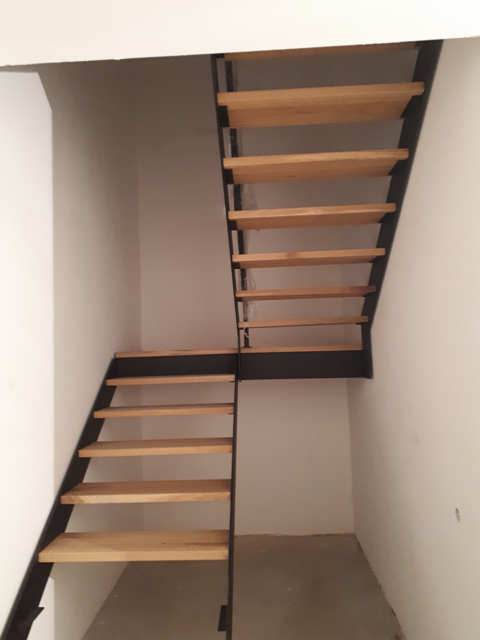 Nowoczesne schody dwubiegowe z drewnianymi stopniami i czarną metalową konstrukcją, umieszczone w surowym wnętrzu z białymi ścianami.