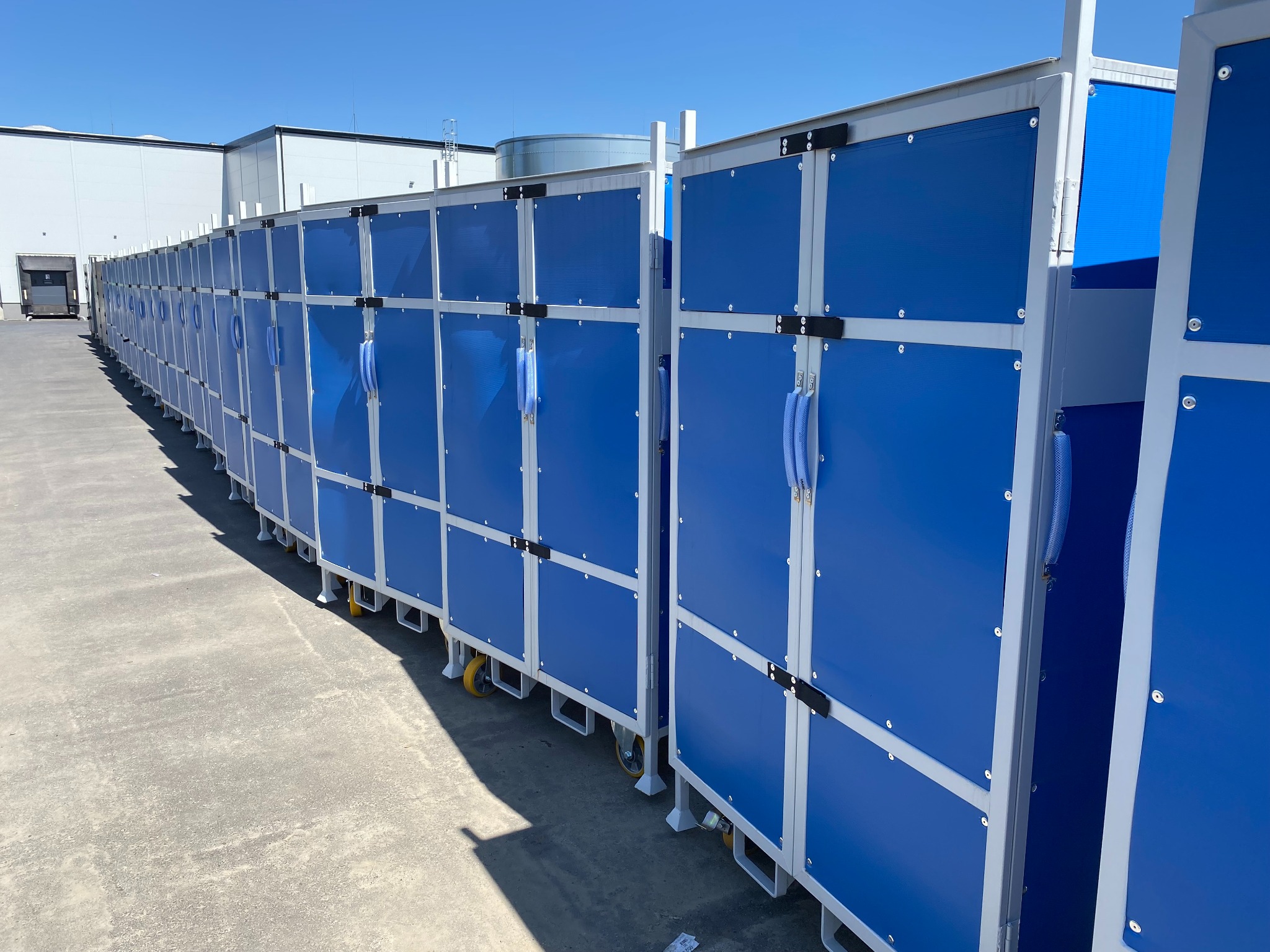 Rząd niebieskich, mobilnych ścianek działowych z metalową ramą i kółkami, ustawionych na zewnątrz budynku przemysłowego w słoneczny dzień.