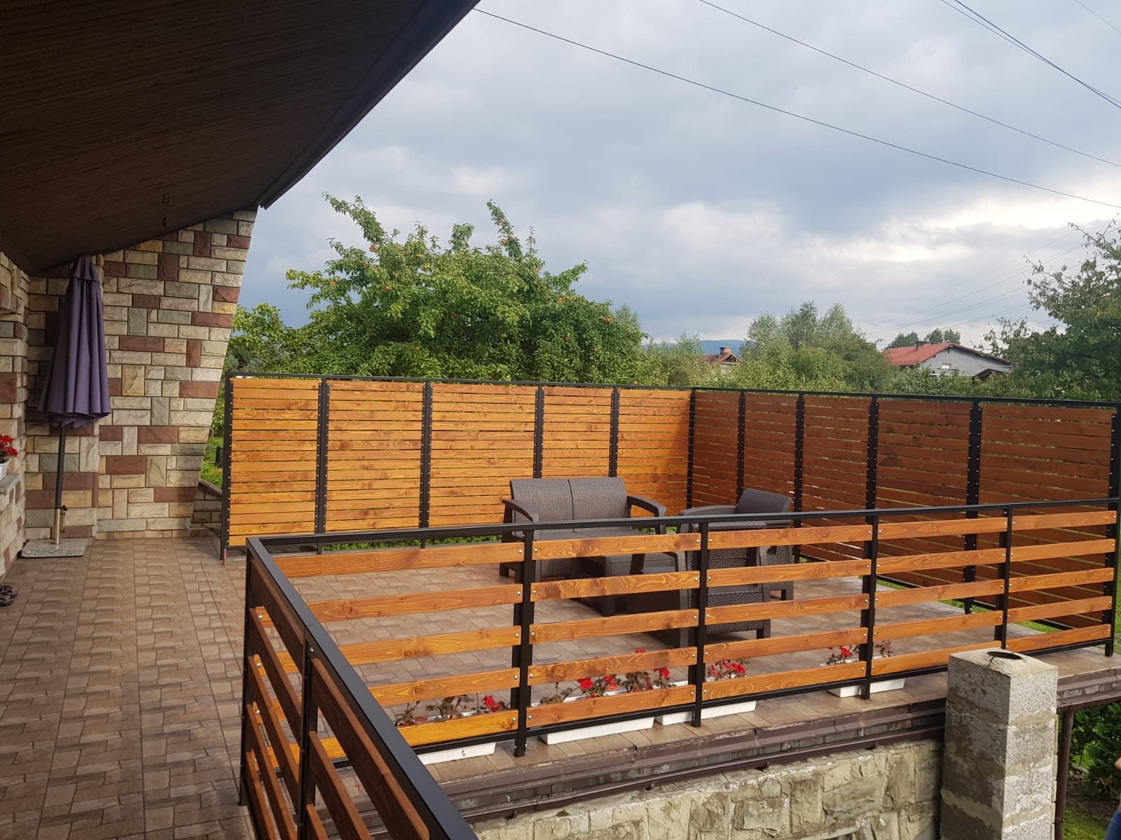 Zadaszony taras z drewnianą podłogą i balustradą z poziomych desek w kolorze miodowego drewna i czarnych metalowych słupków, otoczony drewnianym ogrodzeniem z poziomych desek; w tle zieleń drzew...