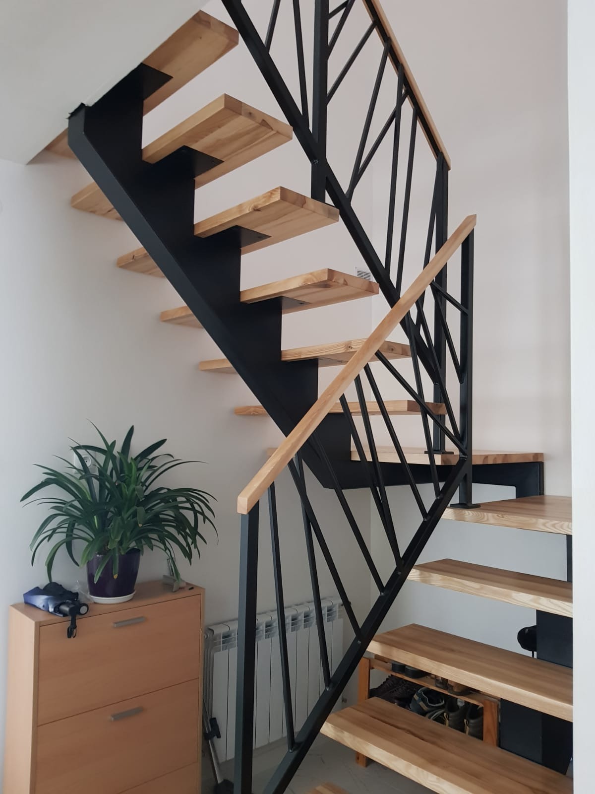 Ażurowe schody z drewnianymi stopniami i czarną, geometryczną balustradą, widok z dołu, obok szafka z rośliną doniczkową.