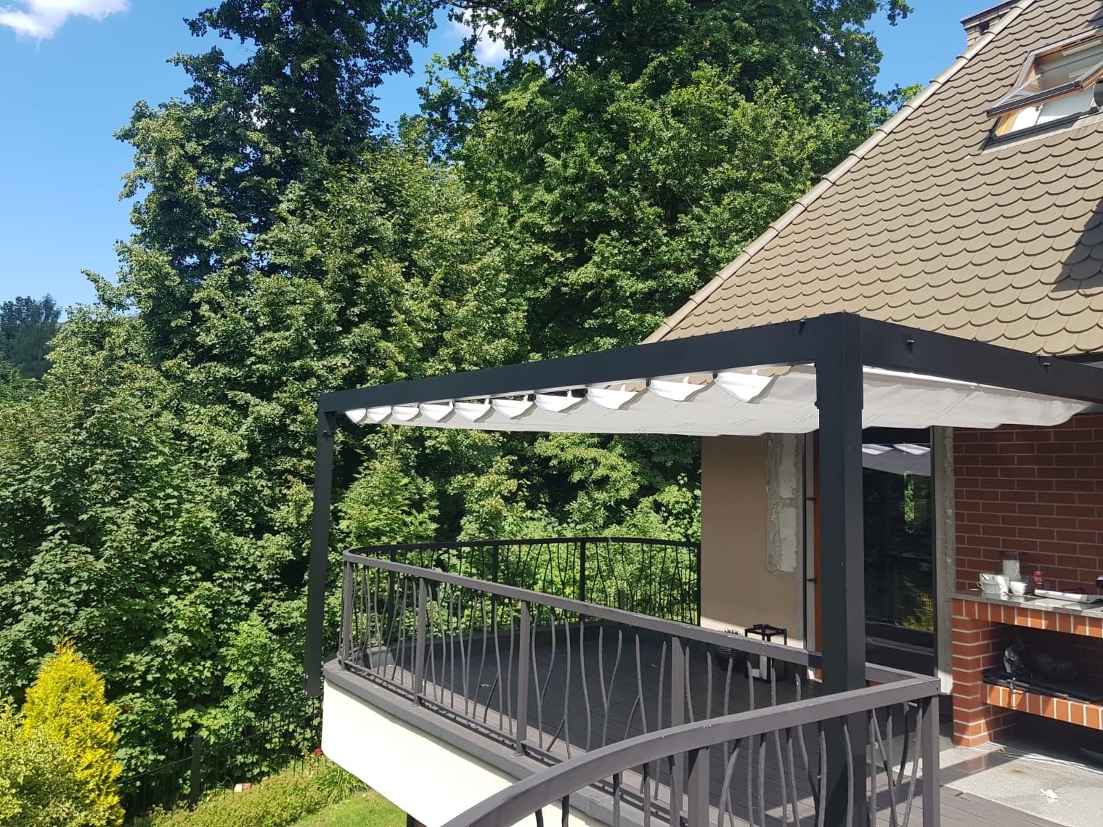 Zadaszony taras z czarną metalową konstrukcją i balustradą, widok na bujną zieleń drzew i dach domu z lukarną.