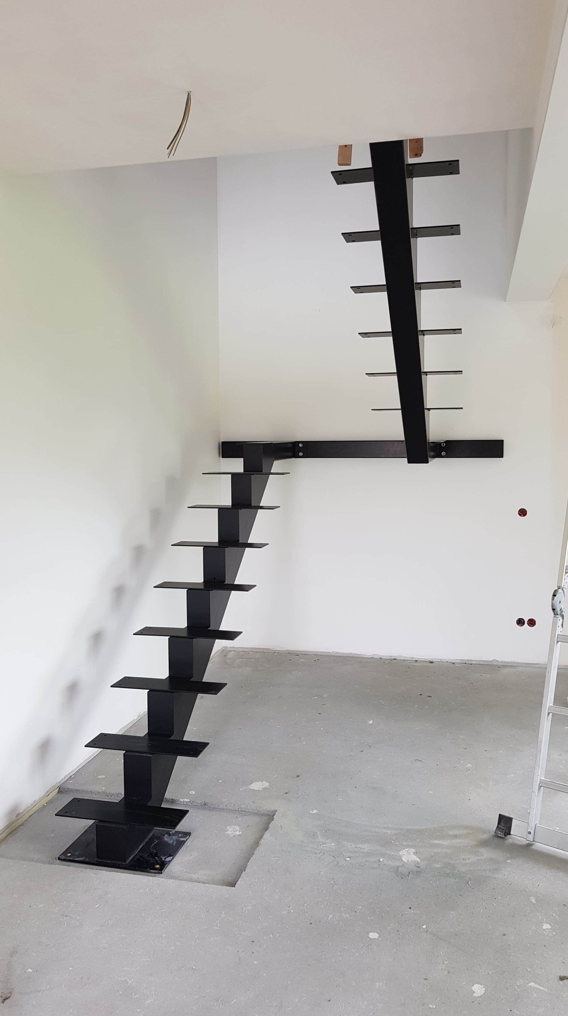 Czarne, minimalistyczne schody wspornikowe z metalu, zamontowane w surowym wnętrzu z betonową podłogą, widoczne otwory w podłodze przygotowane pod konstrukcję.