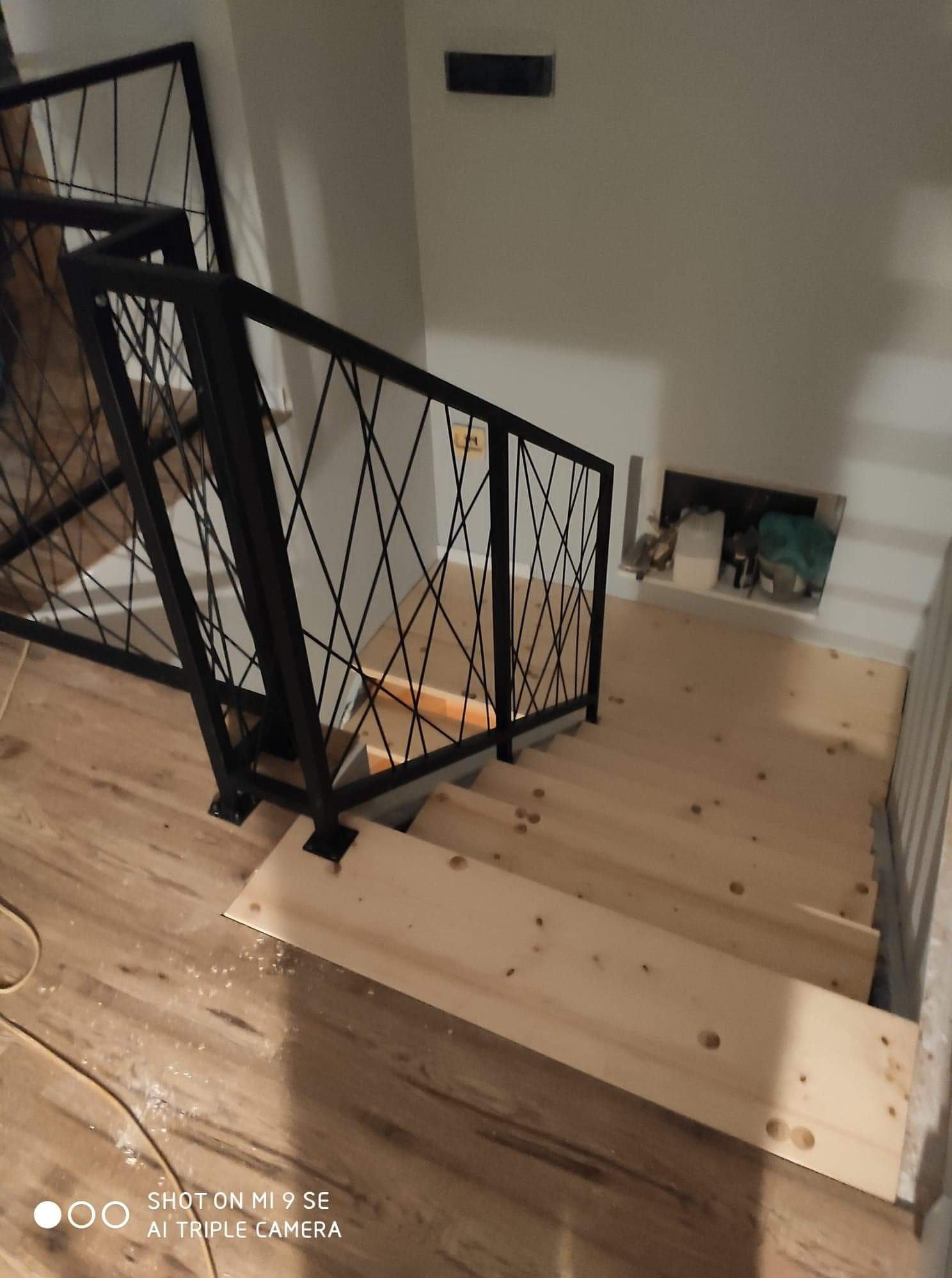 Wnętrze z drewnianymi schodami w trakcie montażu, zabezpieczonymi czarną, metalową balustradą o geometrycznym wzorze z krzyżujących się prętów. Widoczne niedokończone wykończenie i kabel na podłodze.
