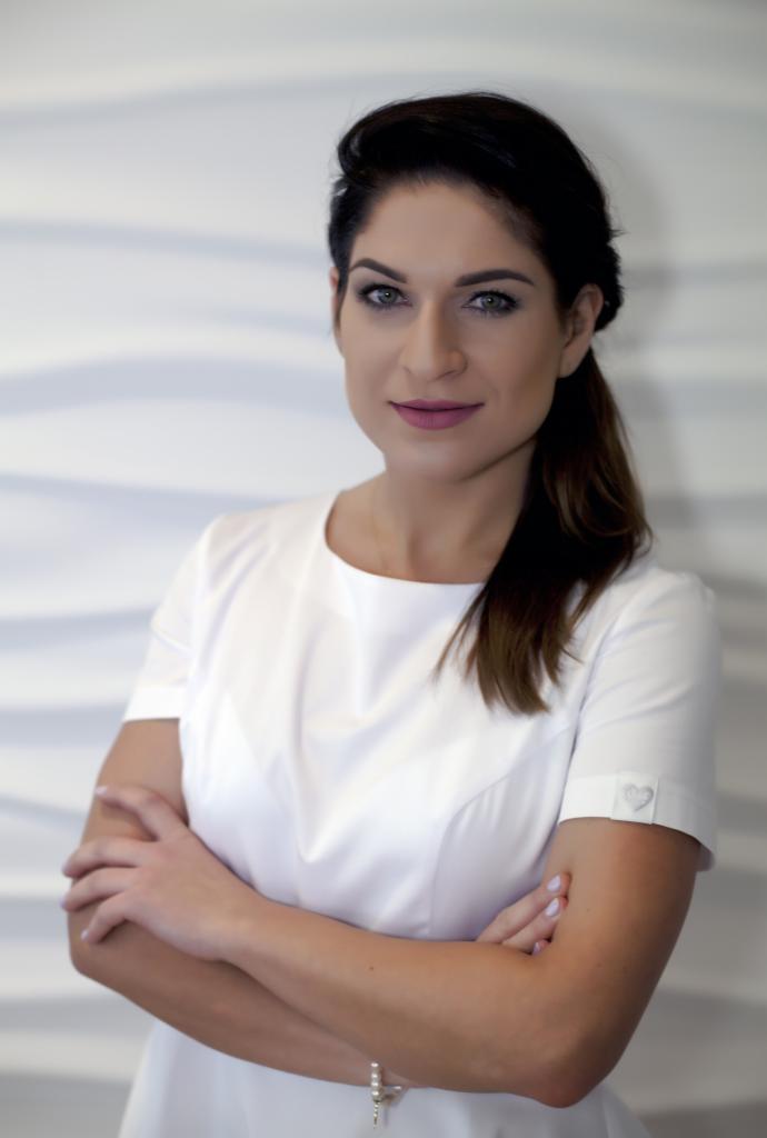 Portret kobiety z ciemnymi włosami, w białym uniformie z haftowanym sercem na rękawie, skrzyżowane ramiona, subtelny makijaż, na tle białej, falistej ściany.