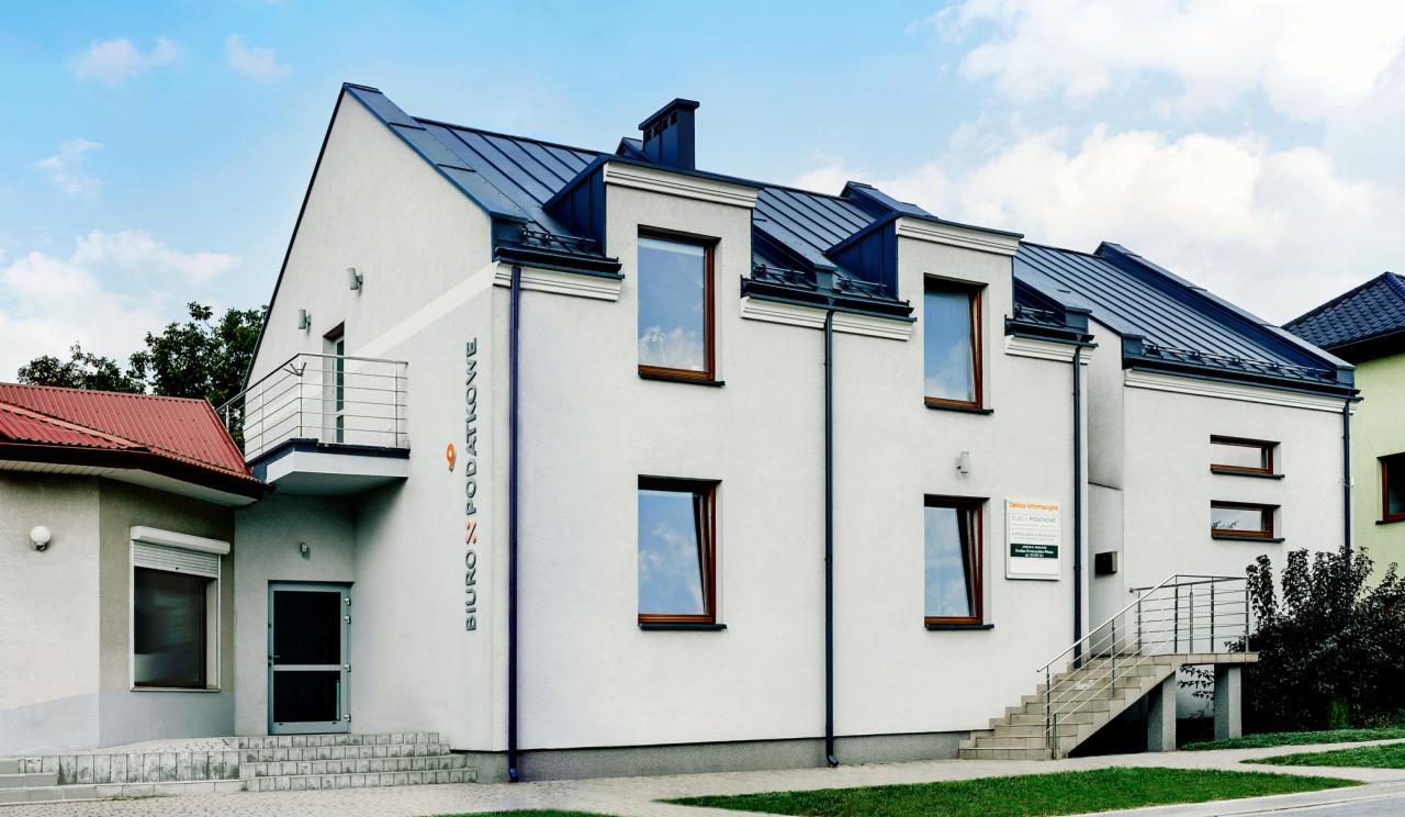 Dwupiętrowy budynek biurowy z szarym dachem i napisem pionowym 'BIURO % PODATKOWE' na elewacji, widok z zewnątrz, z elementami architektury mieszkalnej.