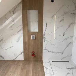 Wykończona łazienka na poddaszu z marmurowymi płytkami i drewnopodobną wnęką z zamontowaną armaturą, widoczny odpływ liniowy i drewnopodobna podłoga.