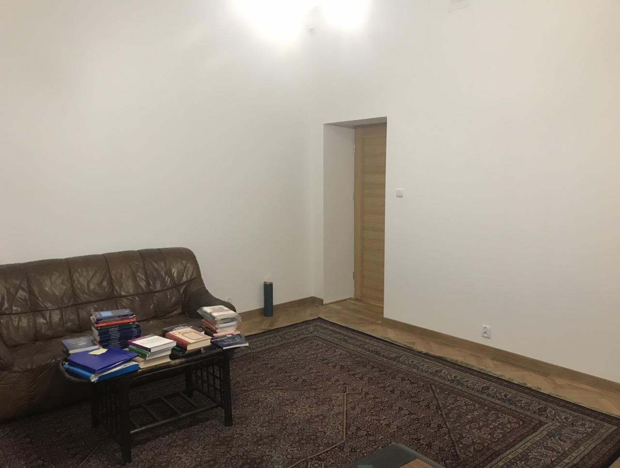 Pokój z brązową skórzaną kanapą, stolikiem kawowym z książkami i dywanem orientalnym przed białymi ścianami i drewnianymi drzwiami.