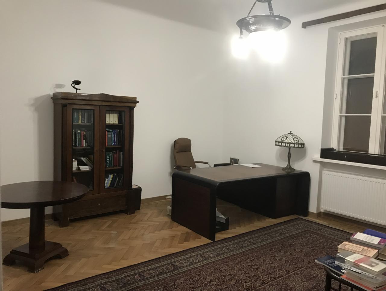 Pokój biurowy z drewnianym biurkiem, biblioteczką z książkami, okrągłym stołem i dywanem w stylu vintage, oświetlony lampą Tiffany i wiszącą lampą sufitową.
