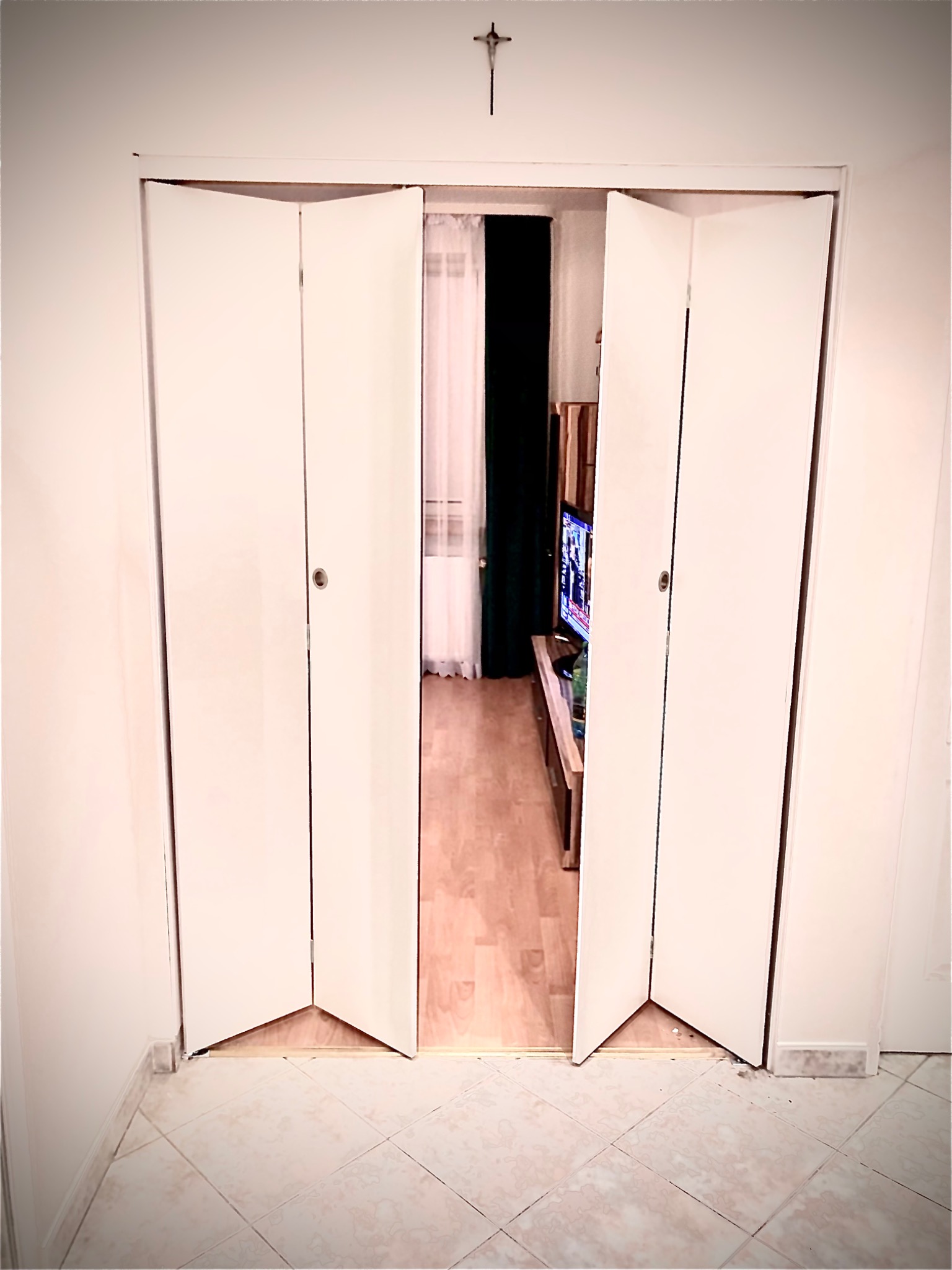 Białe, składane drzwi harmonijkowe, częściowo otwarte, z widokiem na wnętrze pokoju z telewizorem i zasłonami; krzyż na ścianie nad drzwiami.
