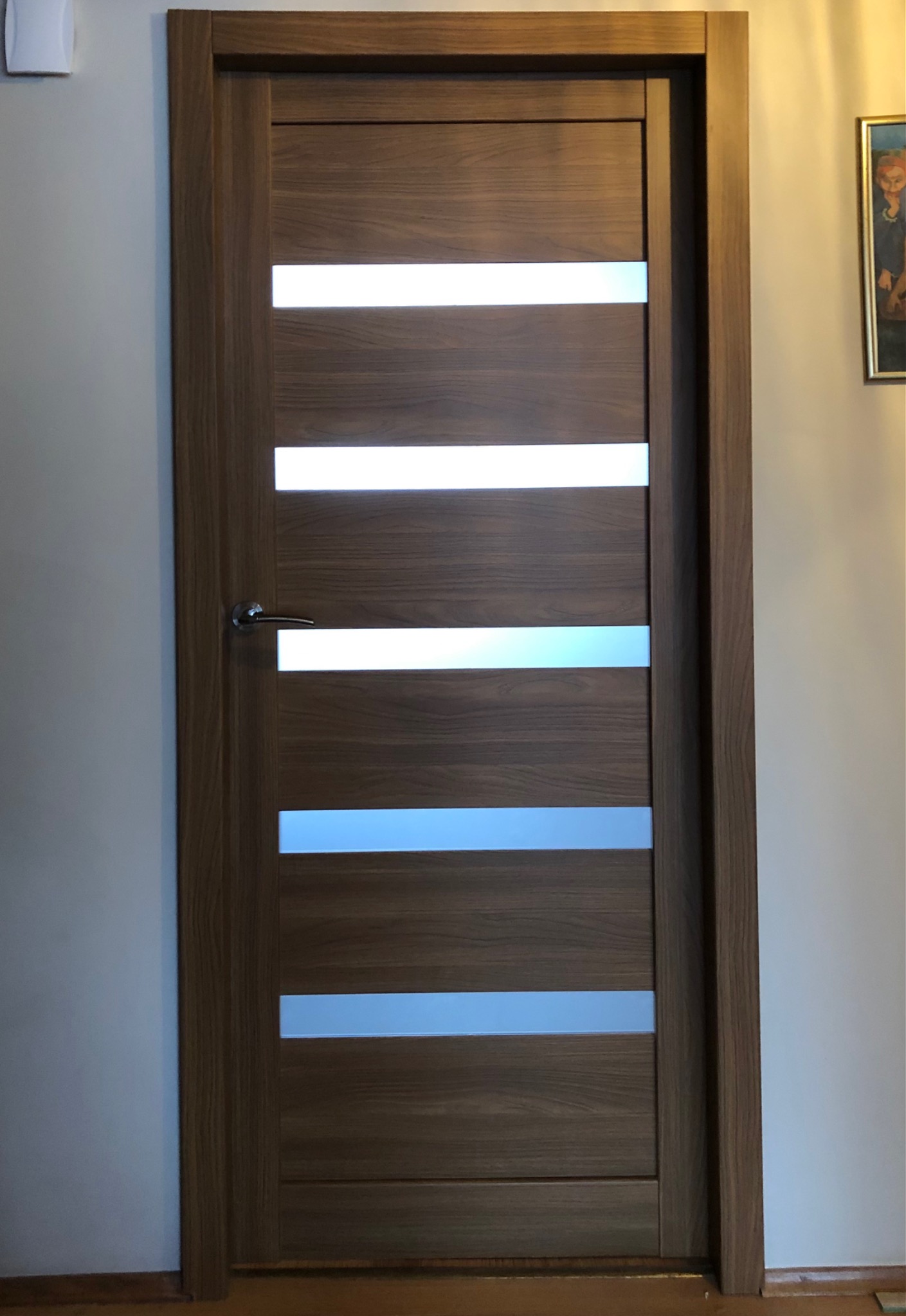Brązowe skrzydło drzwiowe z poziomymi przeszkleniami, widoczna klamka i ościeżnica, na ścianie obraz w złotej ramie.