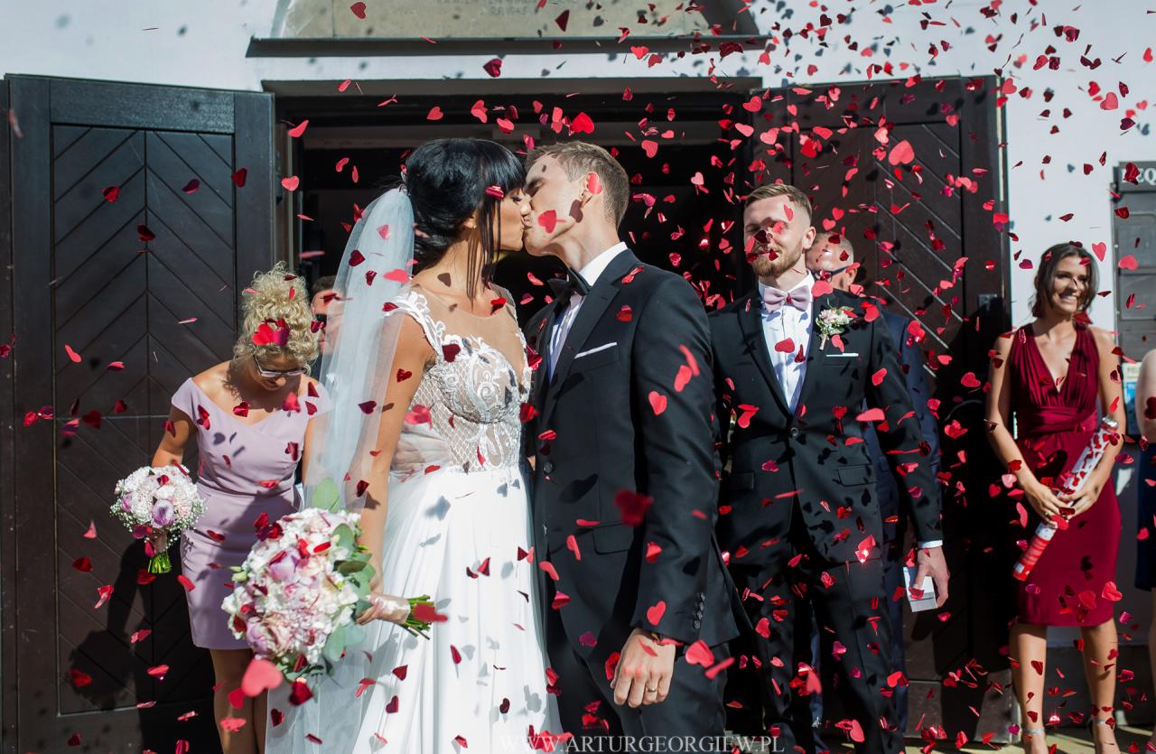 Pocałunek nowożeńców przed kościołem, obsypywanych konfetti w kształcie czerwonych serc, w tle goście weselni i drewniane drzwi.