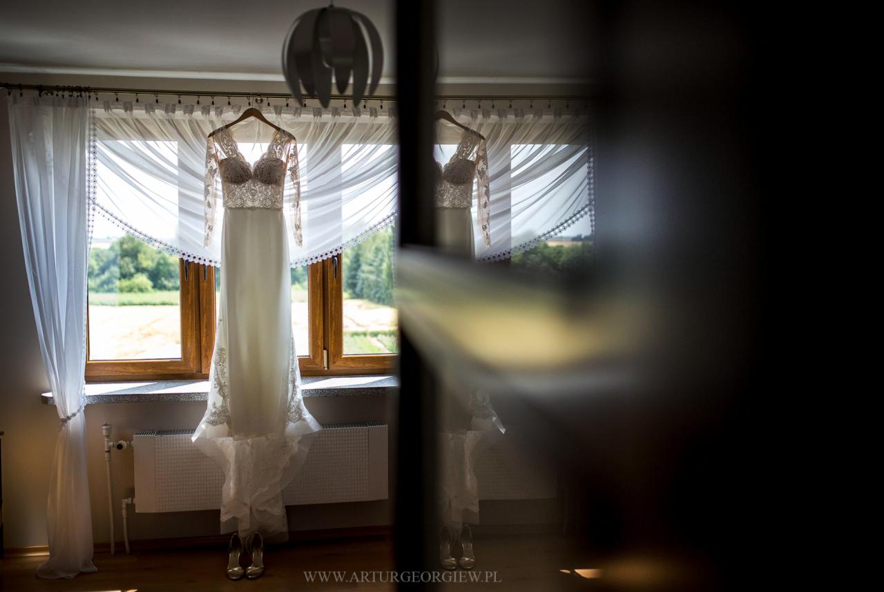 Delikatna suknia ślubna z koronkowymi detalami, wisząca na wieszaku przed oknem z białymi firanami, z widokiem na zielone drzewa w tle, srebrne buty na podłodze, odbicie sukni w szybie po prawej...