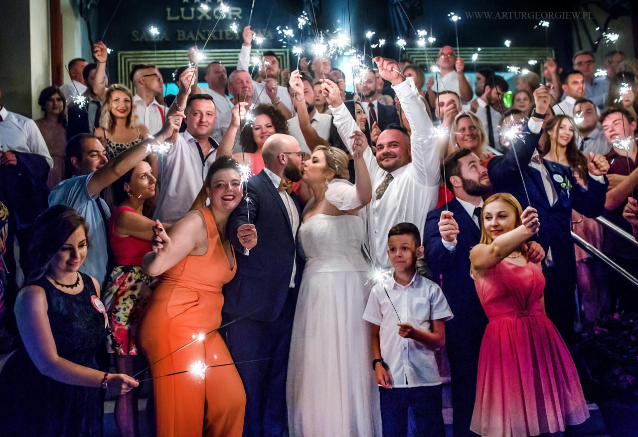 Pocałunek młodej pary otoczonej gośćmi weselnymi z zimnymi ogniami na tle sali bankietowej Luxor.