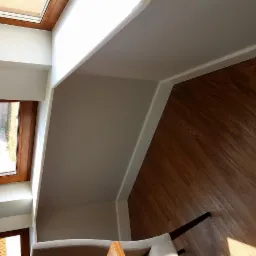 Jasne wnętrze poddasza z oknem dachowym, świeżo pomalowane ściany w odcieniach bieli, panele podłogowe w ciepłym odcieniu drewna, widoczne fragmenty mebli.