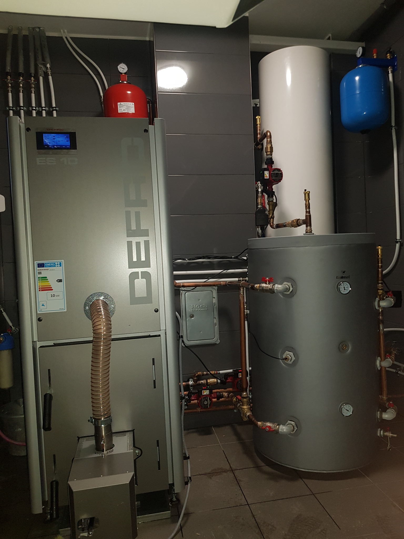 Kotłownia z nowoczesnym piecem Defro ES 10, zbiornikiem akumulacyjnym, naczyniami wzbiorczymi i orurowaniem z miedzi, na tle szarych płytek ceramicznych.