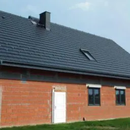 Dom w trakcie budowy z nowym, ciemnoszarym dachem z dachówki, widoczne okno dachowe i komin, ściany z czerwonej cegły, mężczyzna przechodzący obok.