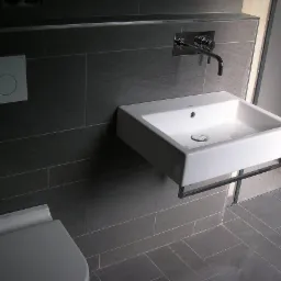 Nowoczesna łazienka z szarymi płytkami, białą umywalką na stelażu i chromowaną baterią ścienną, widoczny fragment toalety.