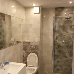 Jasna łazienka z beżowymi i szarymi płytkami, kabiną prysznicową ze szklanymi drzwiami, umywalką z białą szafką i dużym lustrem.