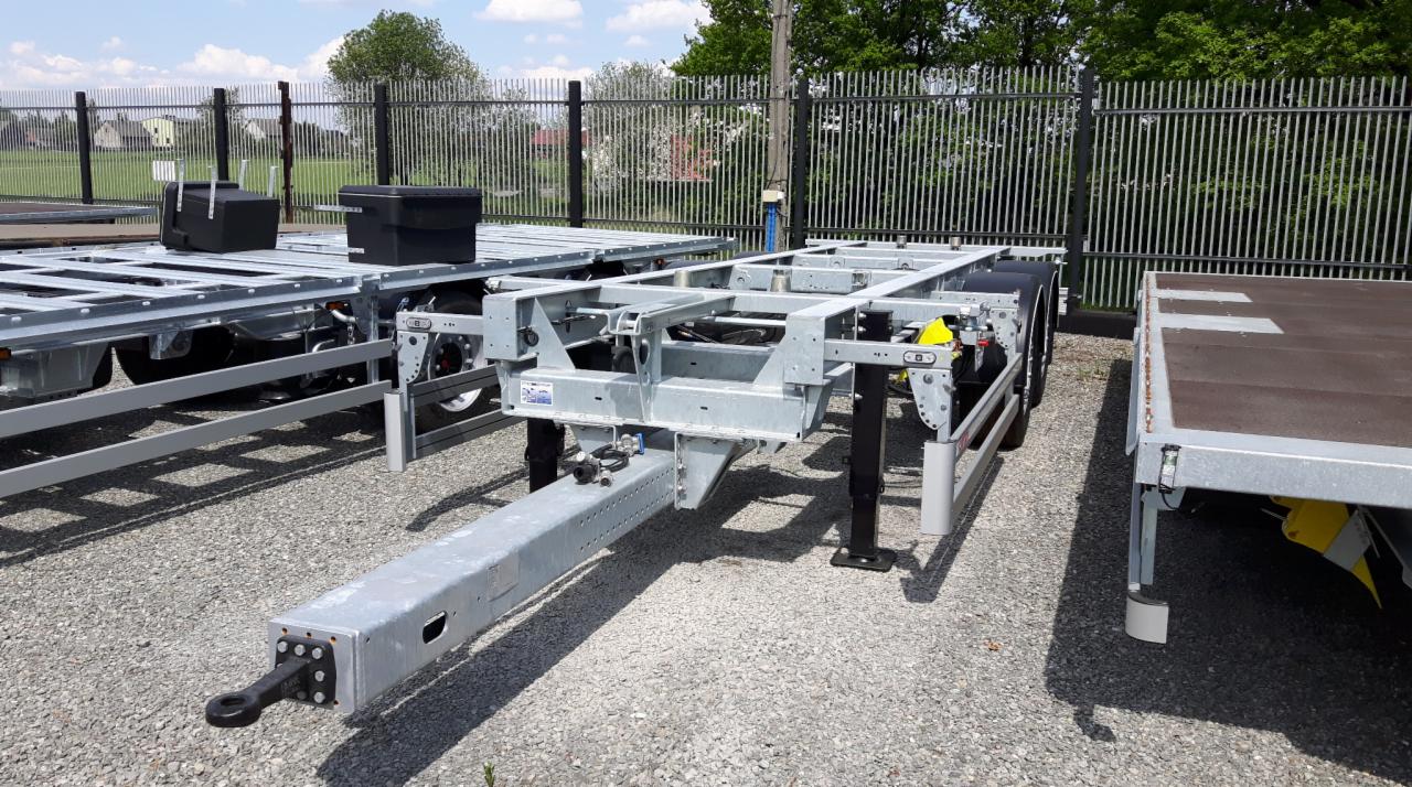 Szara rama naczepy samochodowej z widocznymi kołami i zaczepem, stojąca na żwirowym placu, w tle ogrodzenie i zabudowania.