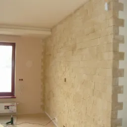 Ściana wykończona beżową płytką imitującą kamień, z widoczną strukturą i fugami, narożnik wykończony wystającymi fragmentami płytek, w tle okno z grzejnikiem i instalacją elektryczną bez osprzętu.