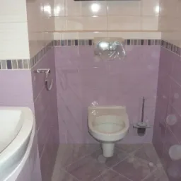 Jasnofioletowa łazienka z beżowymi płytkami u góry, toaleta z deską w kolorze kości słoniowej, plastikowa osłona na spłuczce, okno z drewnianą ramą.