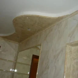 Beżowa łazienka z marmurkowymi płytkami, ozdobnym sufitem podwieszanym z płytek i lustrem w drewnianej ramie.