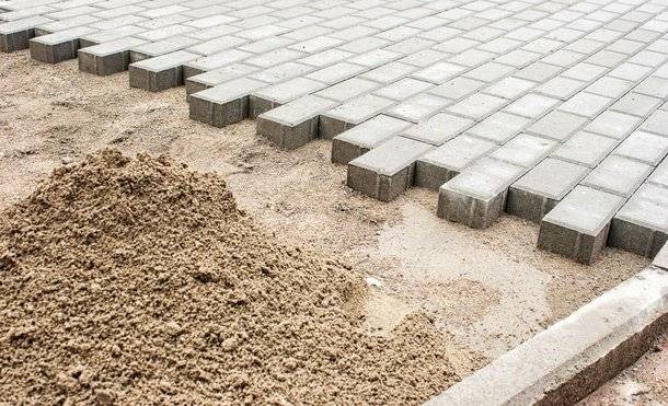 Układanie szarej kostki brukowej na piasku, widoczny fragment krawężnika i usypana góra piasku.