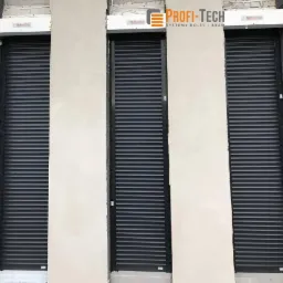 Trzy czarne, częściowo opuszczone systemy osłonowe okienne z widocznym logo producenta na górnej listwie, zamontowane w surowej, nieotynkowanej wnęce okiennej.