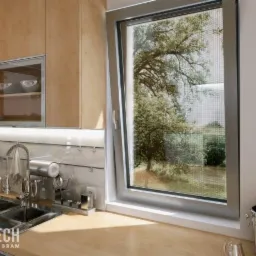 Uchylone okno z zamontowaną siatką przeciw owadom w nowoczesnej kuchni z drewnianymi szafkami i dekoracjami ściennymi.