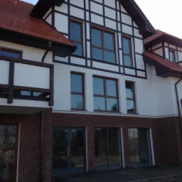 Nowoczesny budynek w Piaskach z wyraźnymi oknami z brązowymi ramami i ceglaną fasadą na parterze, widoczne odbicie drzew w szybach.