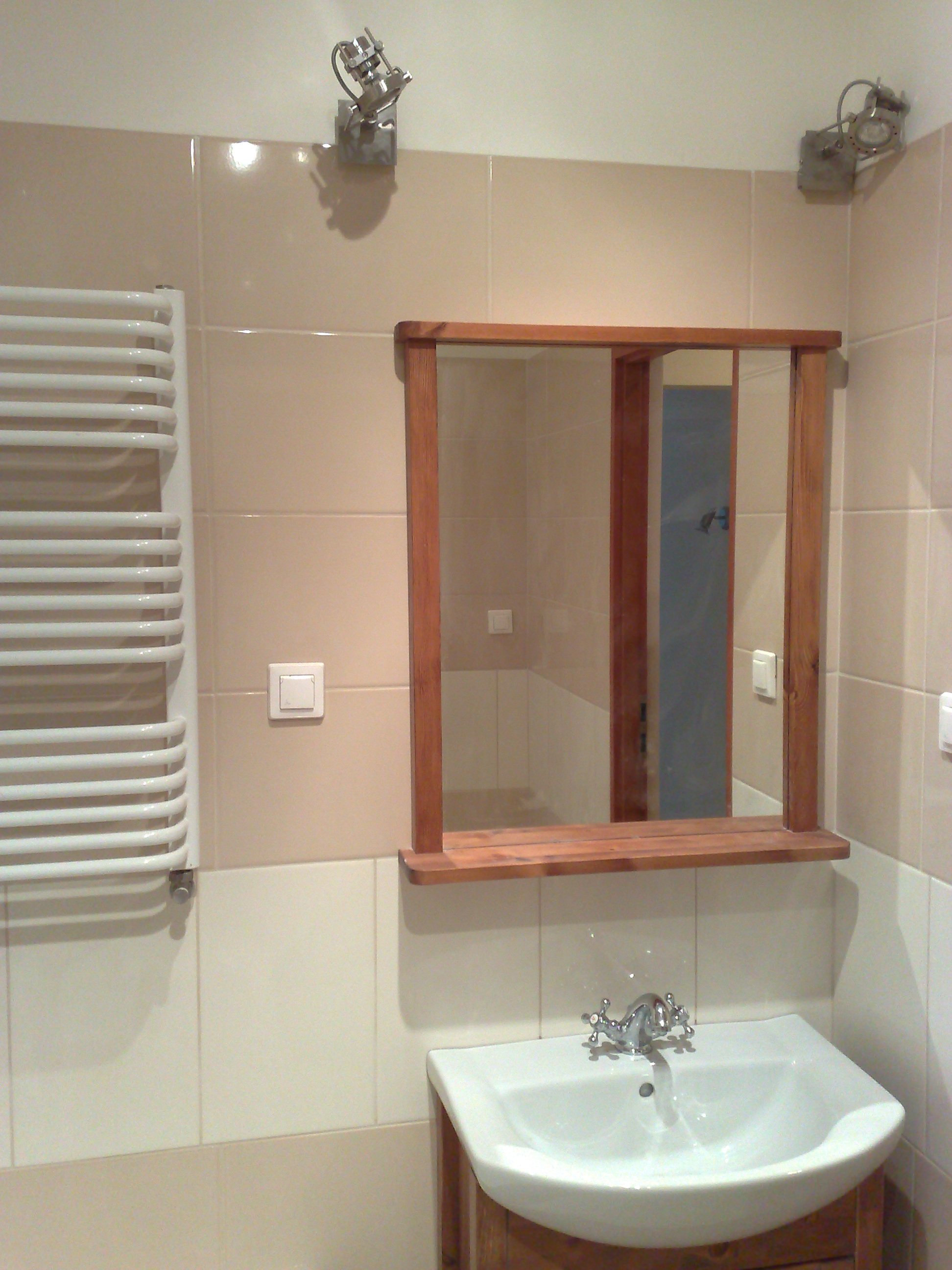 Umywalka z drewnianą szafką i chromowaną baterią, nad nią lustro w drewnianej ramie, na ścianie beżowe płytki i grzejnik drabinkowy.