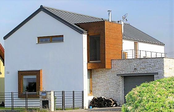 Nowoczesny dom jednorodzinny z białymi ścianami, drewnianą elewacją i kamienną podmurówką, widok z zewnątrz, szara blacha na dachu, metalowe ogrodzenie. Na pierwszym planie stos drewna i zielone...