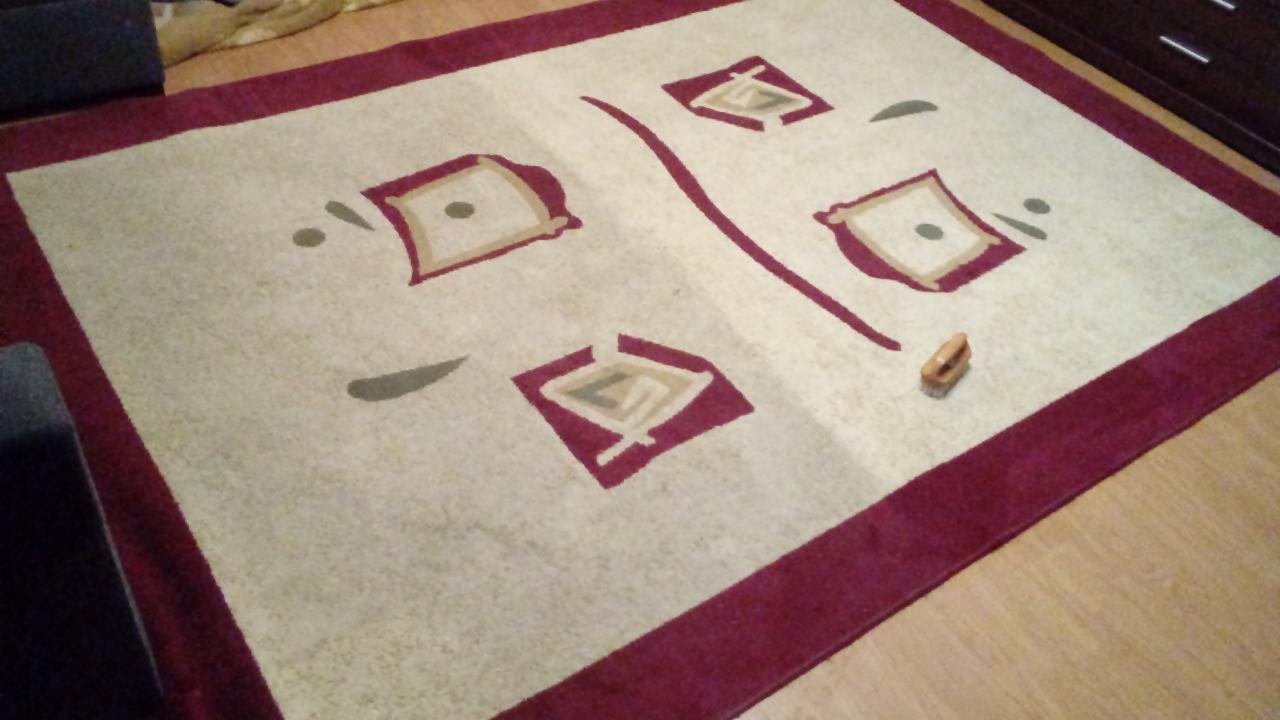 Beżowy dywan z bordową obwódką i geometrycznym wzorem, leżący na drewnianej podłodze, z małą szczotką do czyszczenia obok.