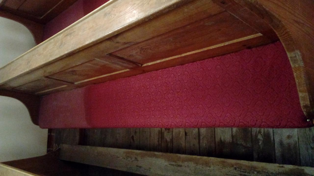 Fragment drewnianej konstrukcji z czerwonym obiciem materiałowym o wzorzystej fakturze, widoczne ślady użytkowania i patyna na drewnie.