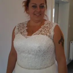 Kobieta w białej sukni z koronkową górą, z tatuażem na ramieniu, uśmiecha się, stojąc w jasnym wnętrzu. Widoczny fragment żyrandola.