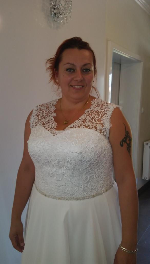 Kobieta w białej sukni z koronkową górą, z tatuażem na ramieniu, uśmiecha się, stojąc w jasnym wnętrzu. Widoczny fragment żyrandola.