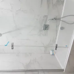 Kabina prysznicowa z przezroczystego szkła z chromowanymi okuciami, zamontowana na ścianie wyłożonej białymi płytkami imitującymi marmur; widoczne niebieskie folie zabezpieczające na szkle...