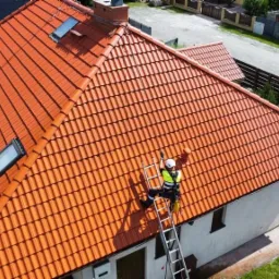 Pracownik w kasku i kamizelce asekuracyjnej na drabinie opiera się o pomarańczowy dach z oknami dachowymi, obok zaparkowany biały samochód dostawczy.