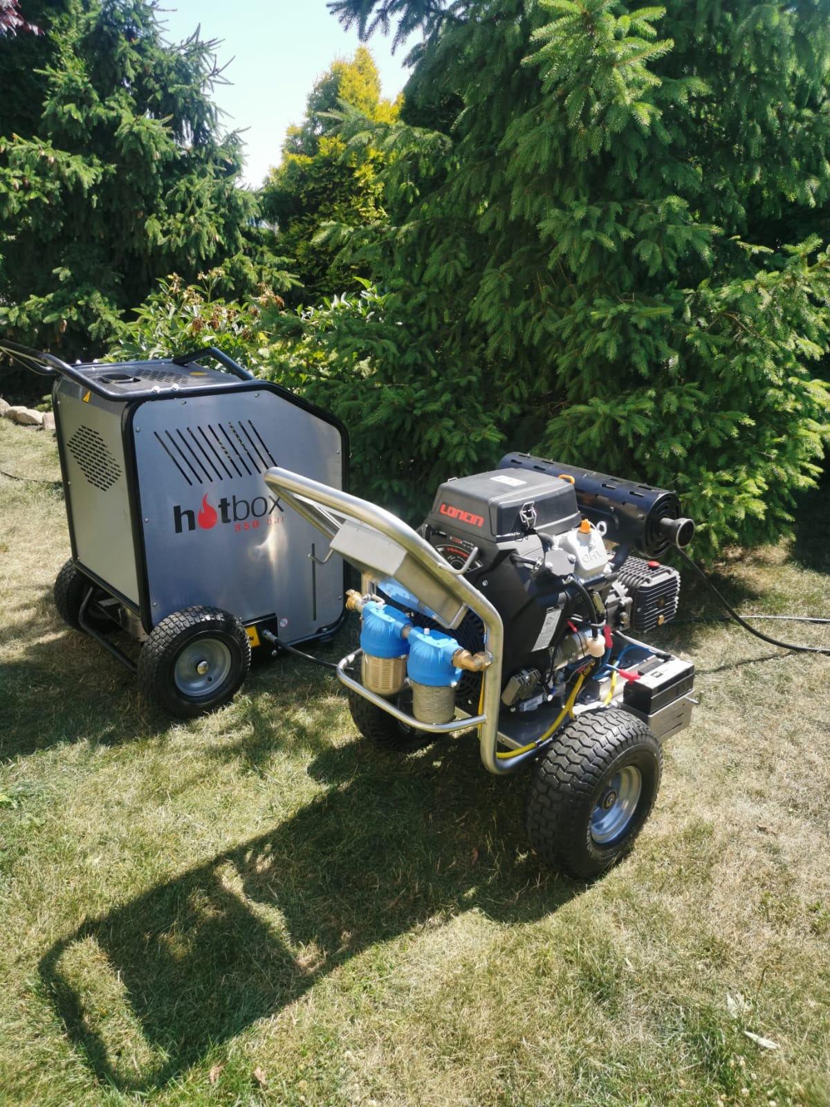 Profesjonalna myjka ciśnieniowa Hotbox 350 bar i Loncin na trawie, otoczona zielenią drzew iglastych, w słoneczny dzień.