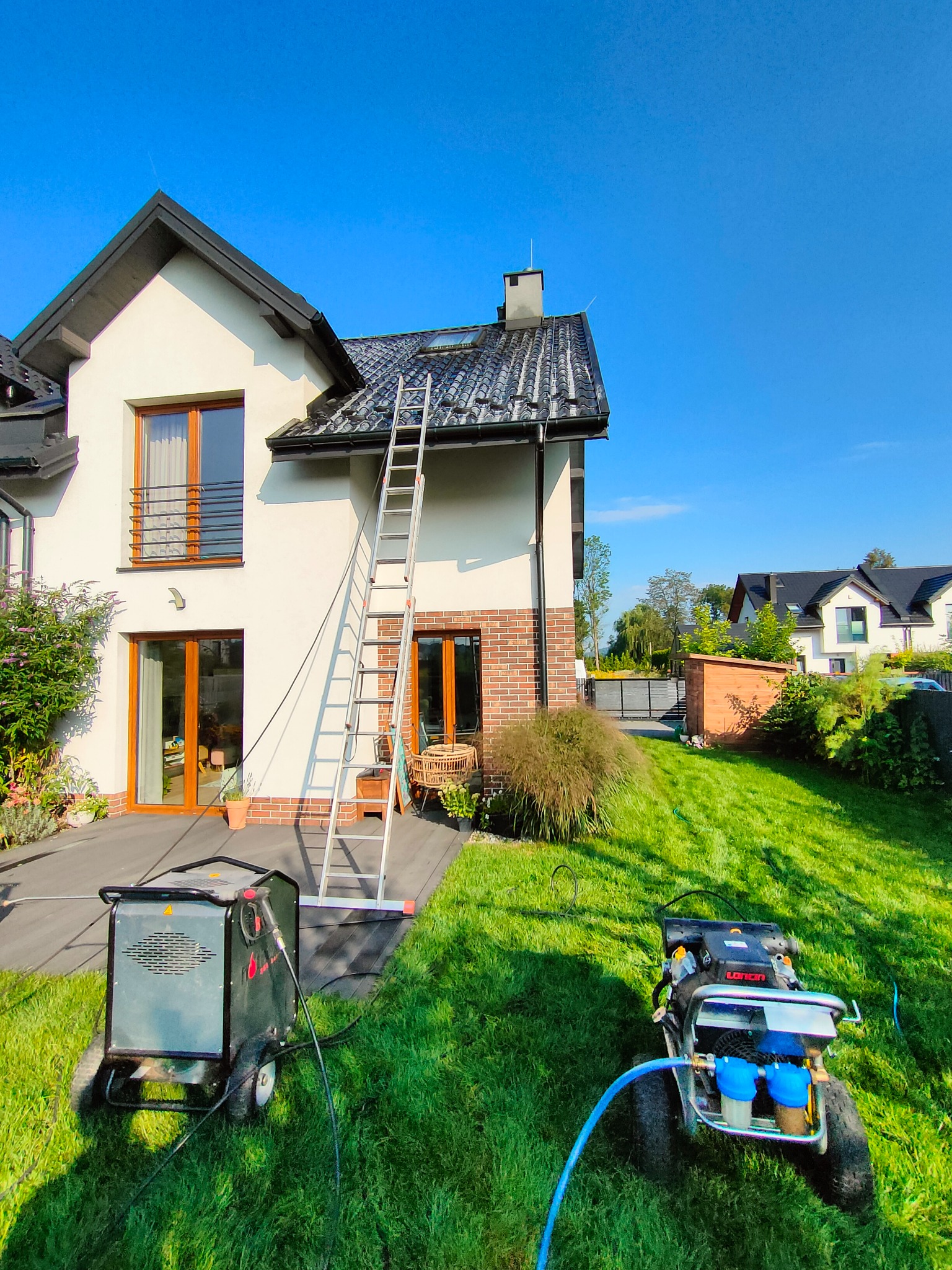 Dwupiętrowy dom z jasną elewacją i ciemnym dachem, z przystawioną drabiną i dwoma urządzeniami wysokociśnieniowymi na trawniku, prawdopodobnie przygotowanymi do czyszczenia.