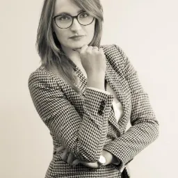 Portret kobiety w okularach i marynarce w pepitkę, z dłonią podpartą pod brodą, w czarno-białej tonacji.